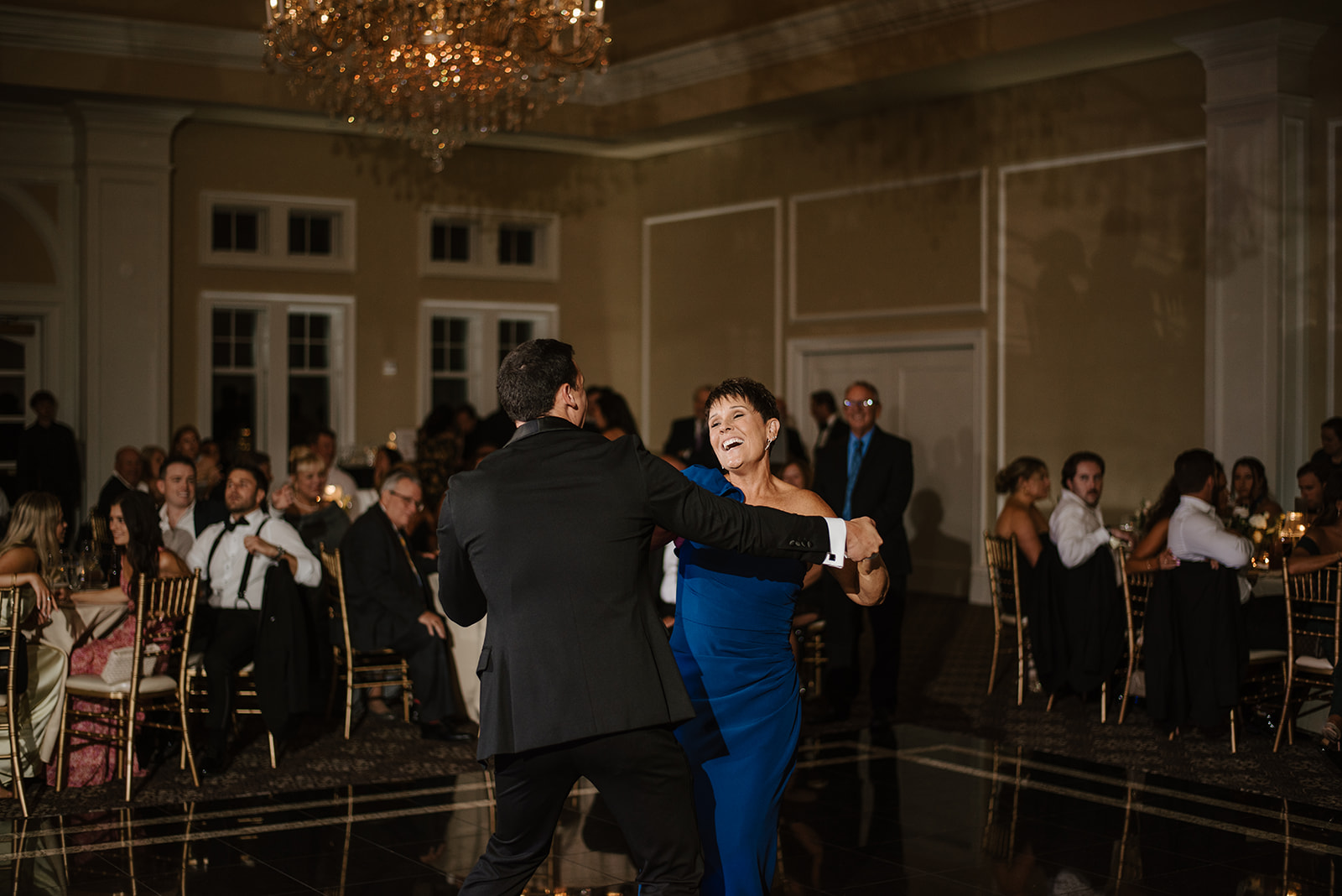 Smiling groom dancing with his laughing mother in a blue gown during the reception mother-son dance.